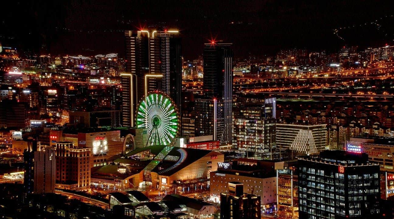 Nighttime cityscape featuring illuminated skyscrapers, a brightly lit Ferris wheel, and a busy urban area.