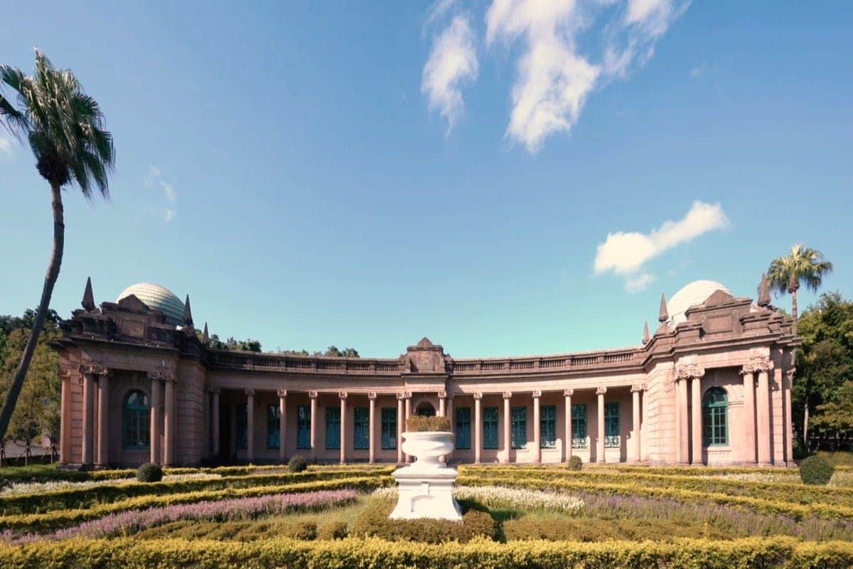 A neoclassical building with columns and domes, surrounded by neatly trimmed gardens and palm trees under a blue sky.
