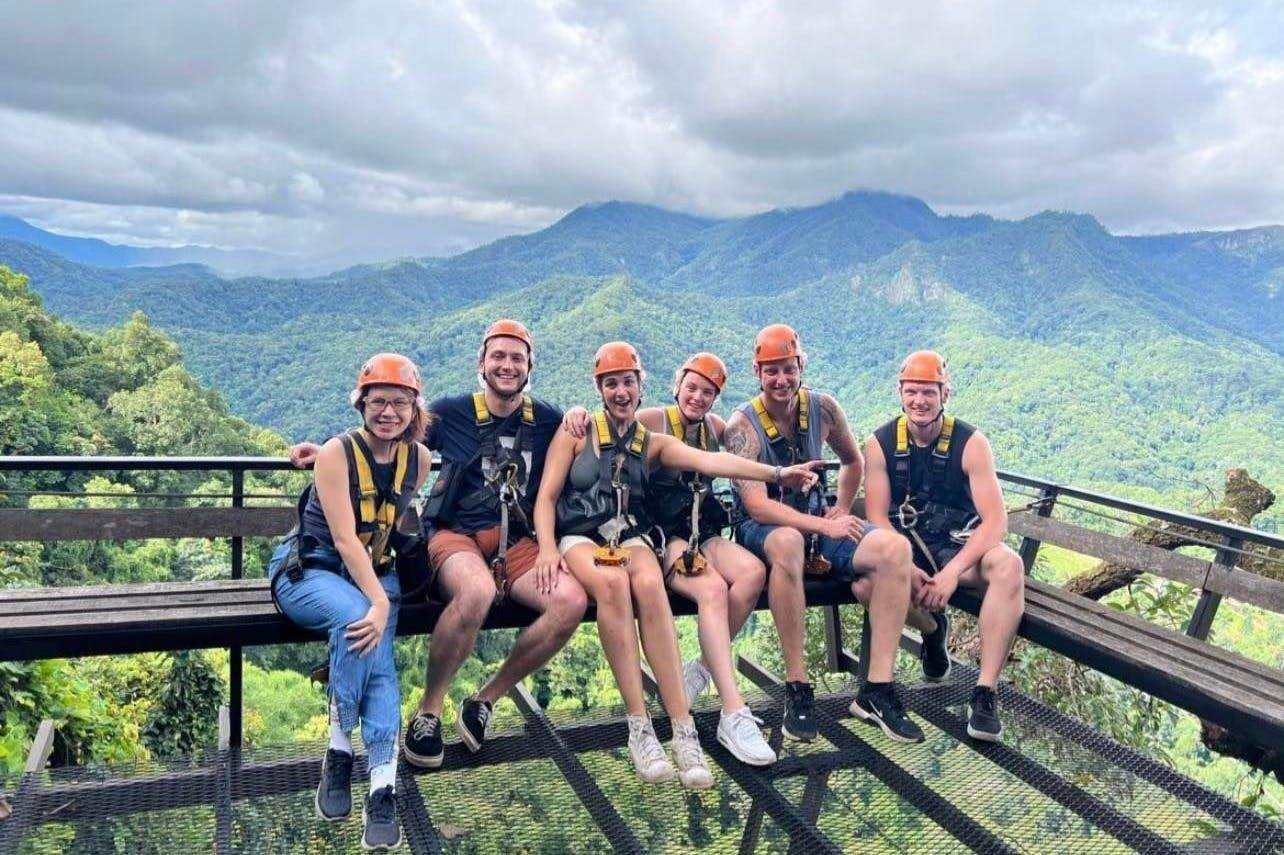 Six people in harnesses and helmets sit on a platform with a lush, mountainous forest in the background.