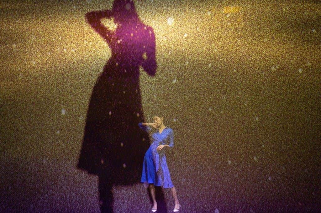 A person in a blue dress poses dramatically against a textured backdrop, with their shadow projected behind them.