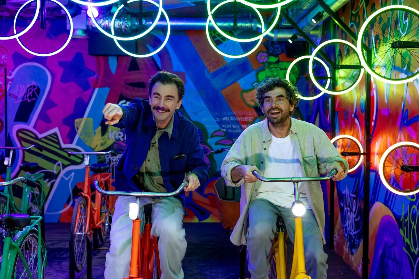 Two men riding colorful bicycles in a vibrant, neon-lit space with graffiti walls and illuminated bicycle wheels overhead.