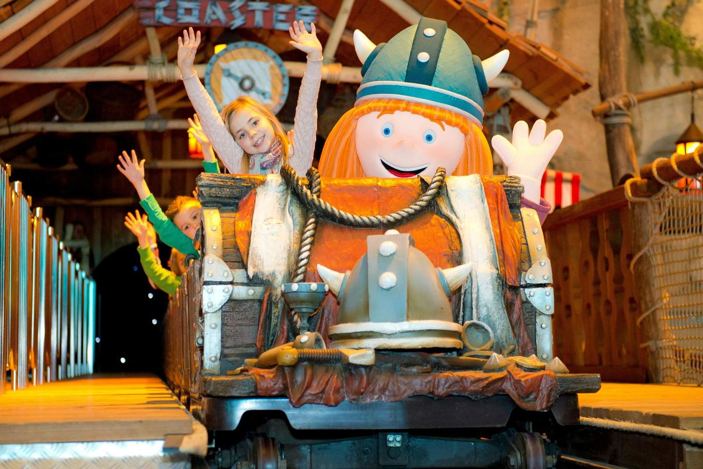 Children with raised hands on an amusement ride featuring a cartoon Viking theme, with wooden decor and festive lighting.