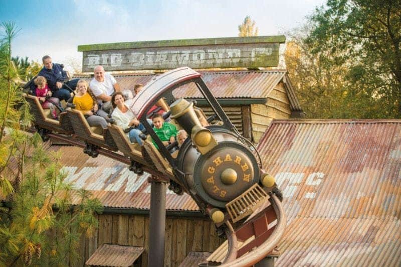 People riding a train-themed roller coaster in front of a rustic, wooden building on a sunny day.