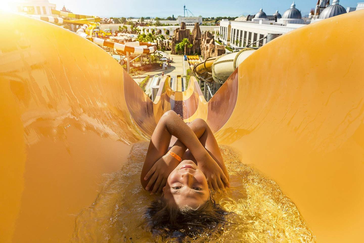 Person sliding down a yellow waterslide, crossing their arms over their chest, with an amusement park in the background.