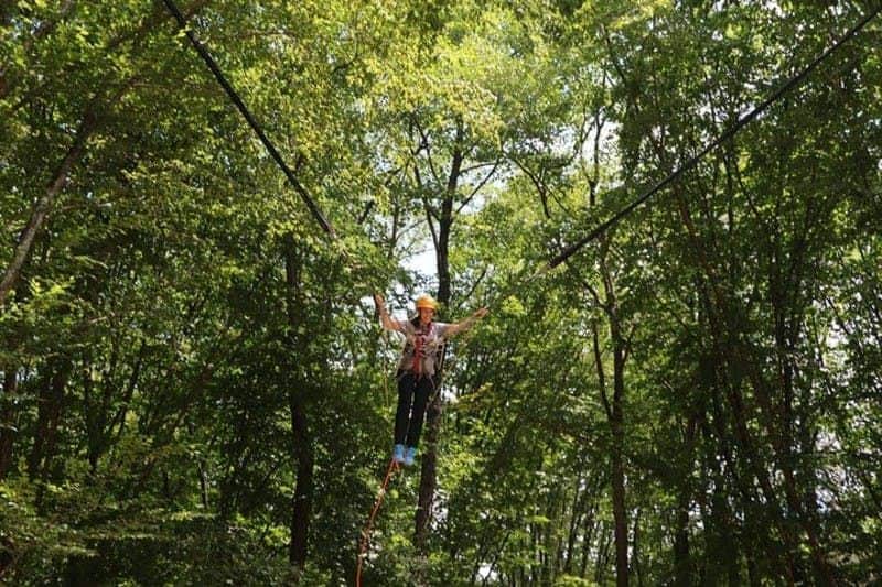 Forest Kemerburgaz A person with a helmet and harness balances on a rope between trees in a lush forest environment.