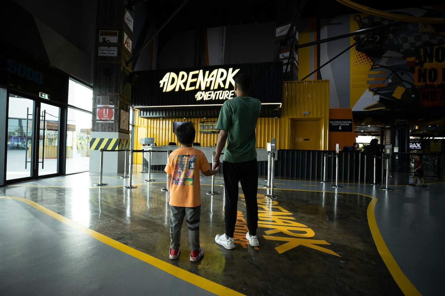Adrenark Adventure A child and an adult stand hand in hand, facing the entrance to "Adrenark Adventure," illuminated by a neon sign.