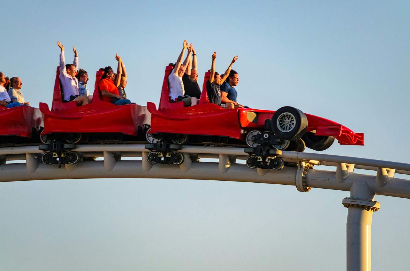 People with raised hands riding in red roller coaster cars shaped like race cars, under a clear blue sky.