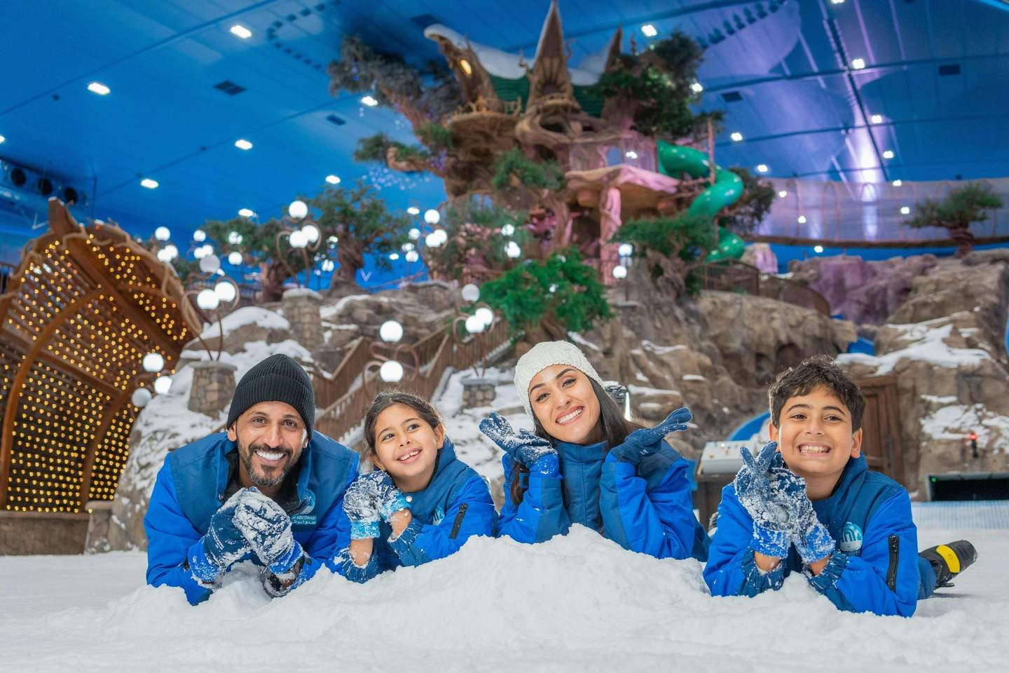 Snow Abu Dhabi A family of four in winter attire lies on snow in a decorated indoor park, smiling and posing for the camera.
