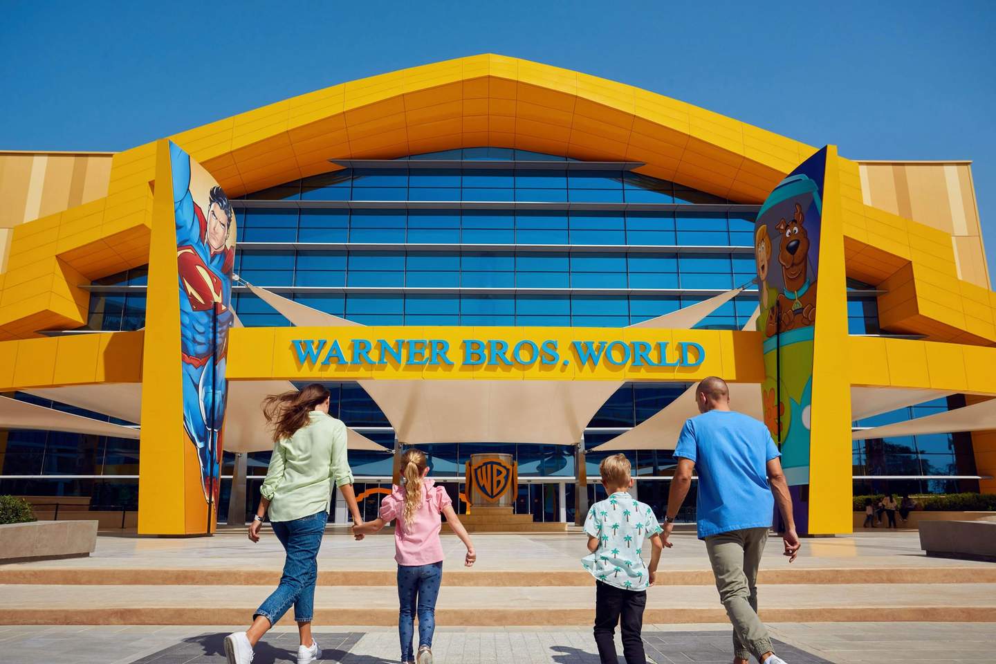 A family walking towards the entrance of Warner Bros. World, with Superman and Scooby-Doo artwork on the building's facade.