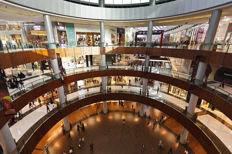 A spacious multi-level shopping mall with numerous stores and people walking along the glass-railed walkways.