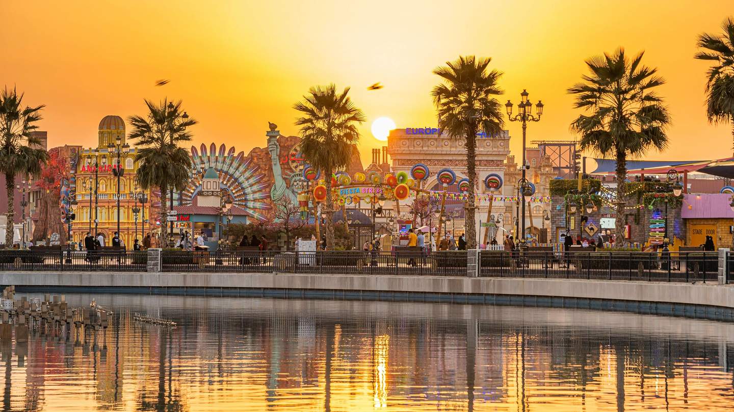 Amusement park at sunset with colorful structures, palm trees, and people walking along a waterfront. Reflections in the water.