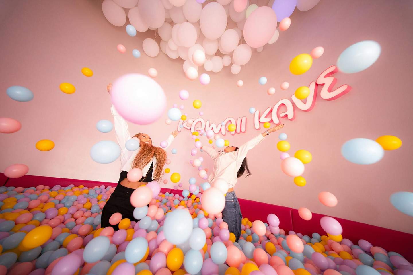 Two people joyfully throw pastel balloons in a colorful ball pit, under hanging white balloons, with the sign "KAWAII KAVE" on the wall.