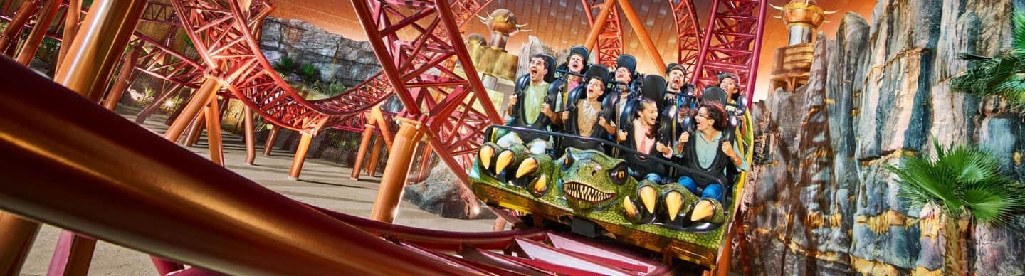 People riding a rollercoaster with a dinosaur-themed front car, surrounded by a red track structure and artificial rock formations.