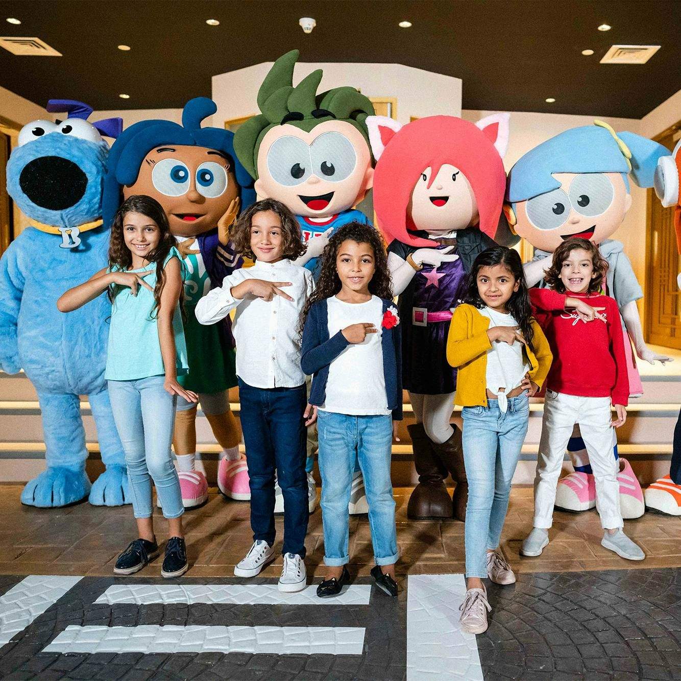 Five children stand in front of large colorful character mascots, smiling and pointing, under indoor lighting.