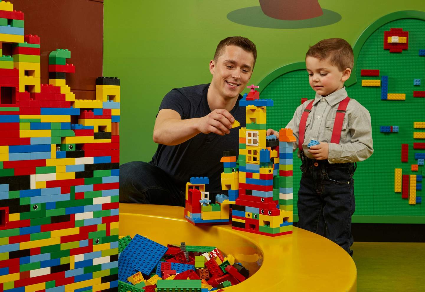 An adult and a child building colorful LEGO structures on a yellow table with a green background.