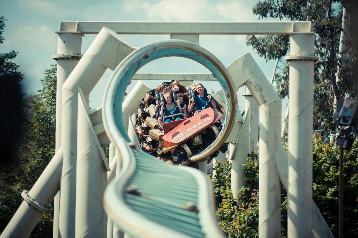 Smiling people riding a roller coaster through a loop on a sunny day, surrounded by trees and white structural supports.