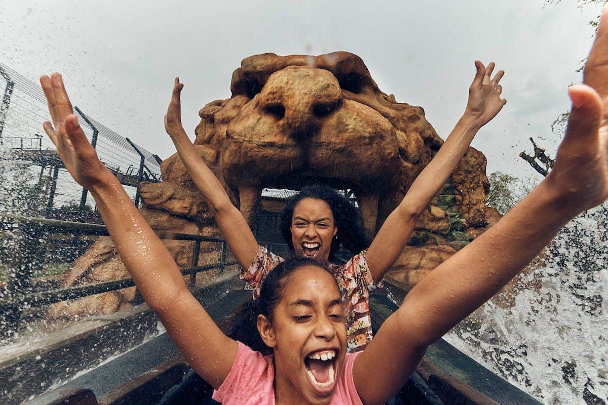Two people on a water ride, arms raised, screaming with joy as they pass under a large rock formation.