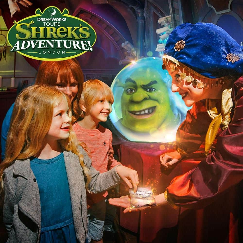 A woman and two children interact with a costumed character next to a crystal ball displaying Shrek’s face at Shrek’s Adventure! London.