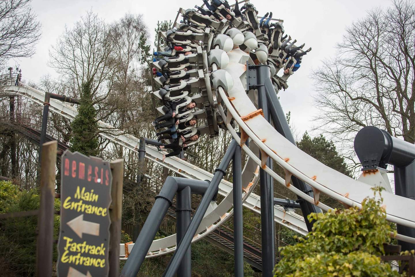 People riding an inverted rollercoaster, heads downward, with a "Main Entrance" and "Fastrack Entrance" sign in the foreground.