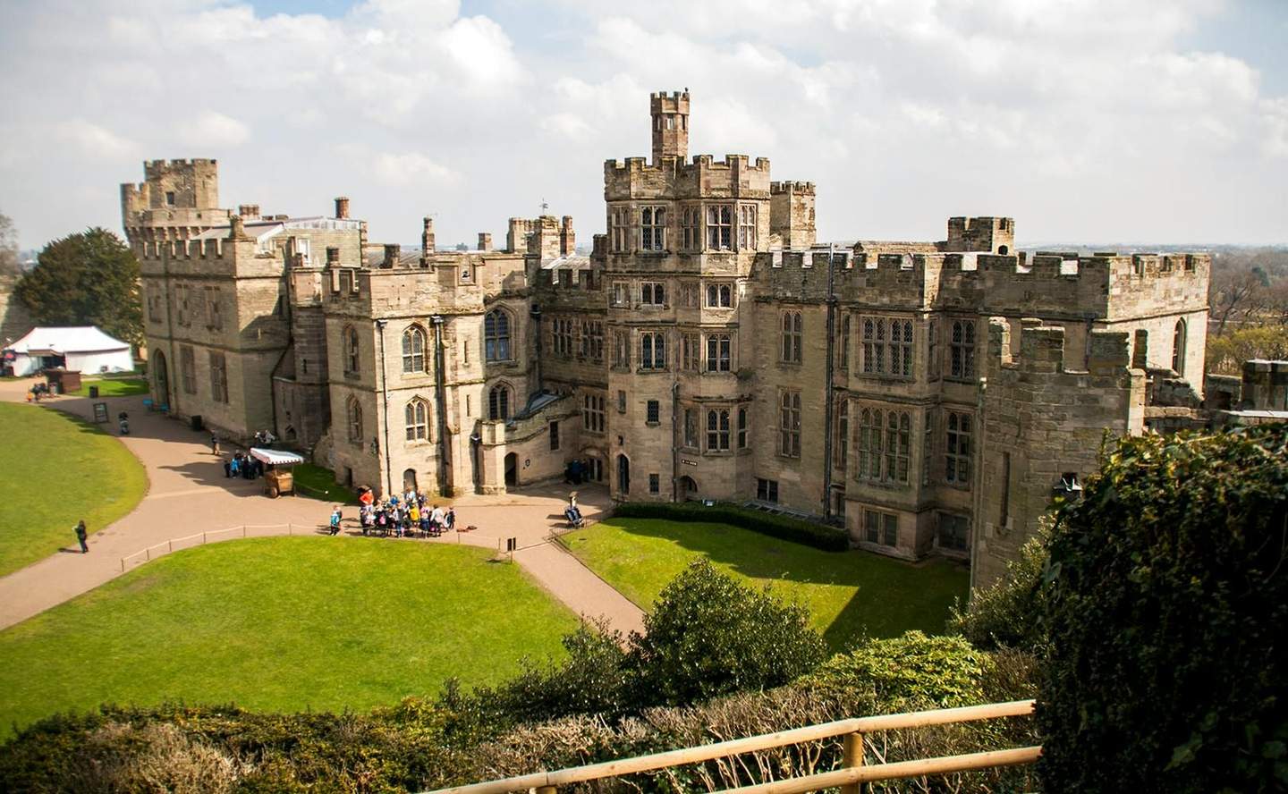 Château de Warwick Grand château de pierre avec de multiples tours et fenêtres, entouré de pelouses et d'allées. Des personnes sont rassemblées près de l'entrée.