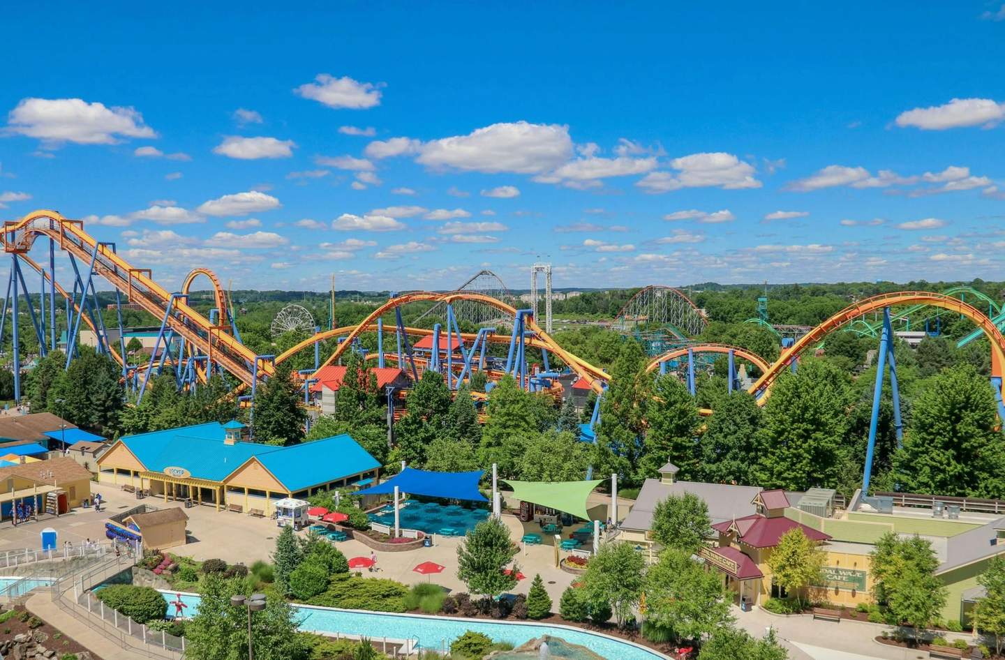 A theme park with colorful roller coasters, green trees, buildings, seating areas, and a bright blue sky with scattered clouds.