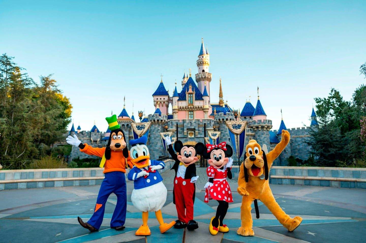 Disney characters stand in front of a castle with blue and gold turrets, set against a clear sky and flanked by greenery.
