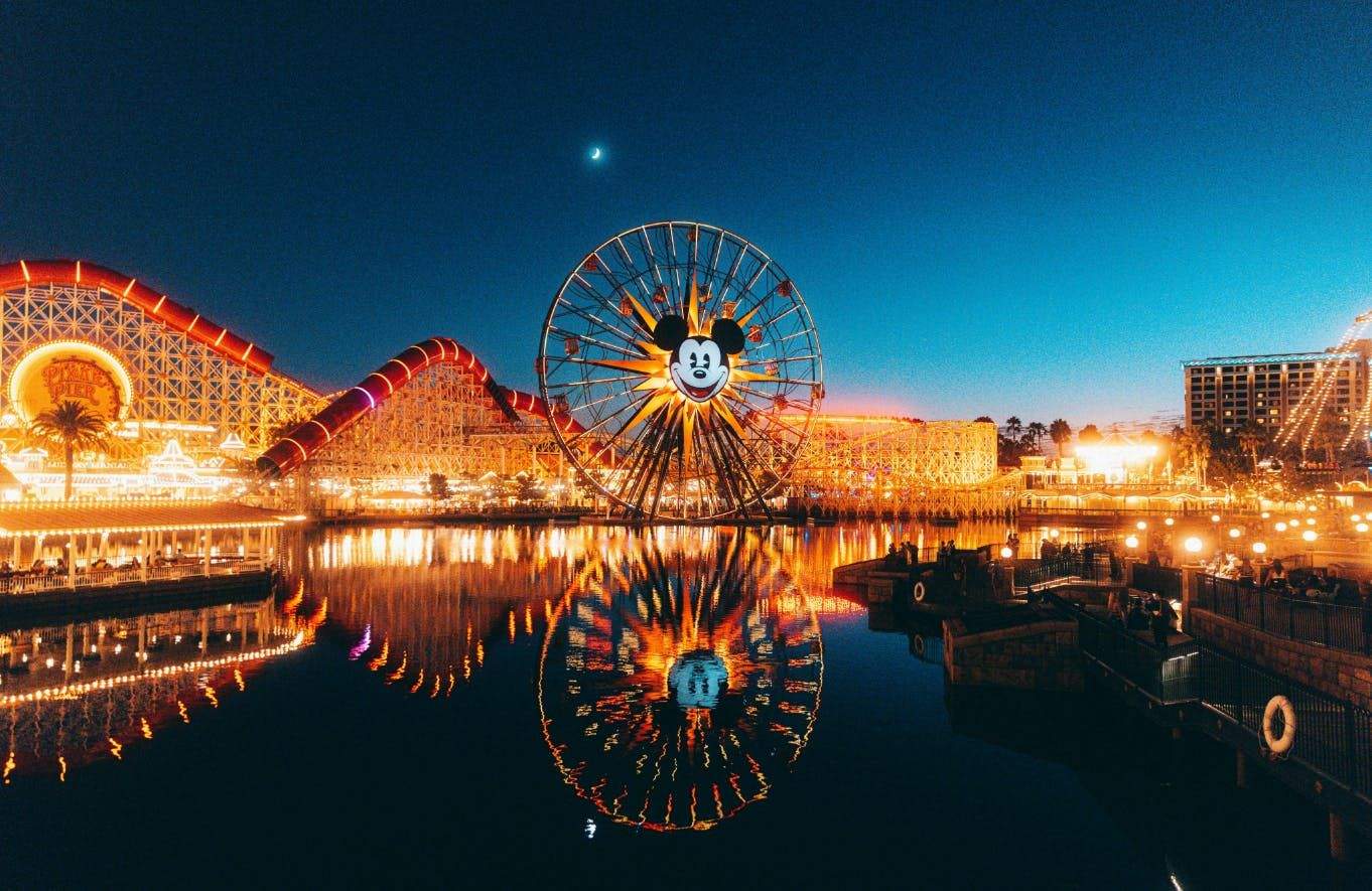 Amusement park at dusk with a lit Ferris wheel featuring a Mickey Mouse design, roller coaster, and waterfront reflections.