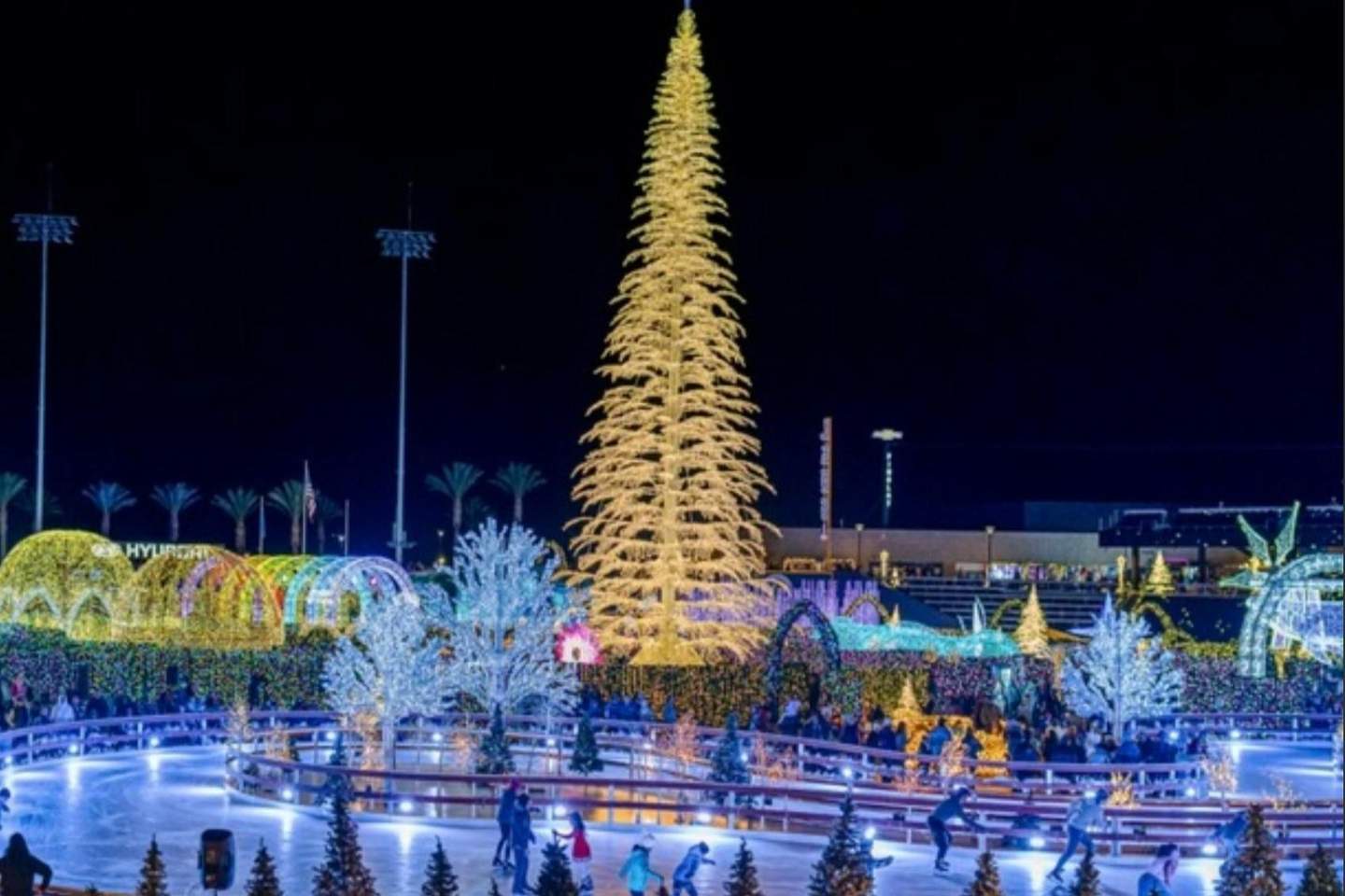 A brightly lit outdoor ice skating rink surrounded by festive holiday lights and decorations, including a large illuminated tree.