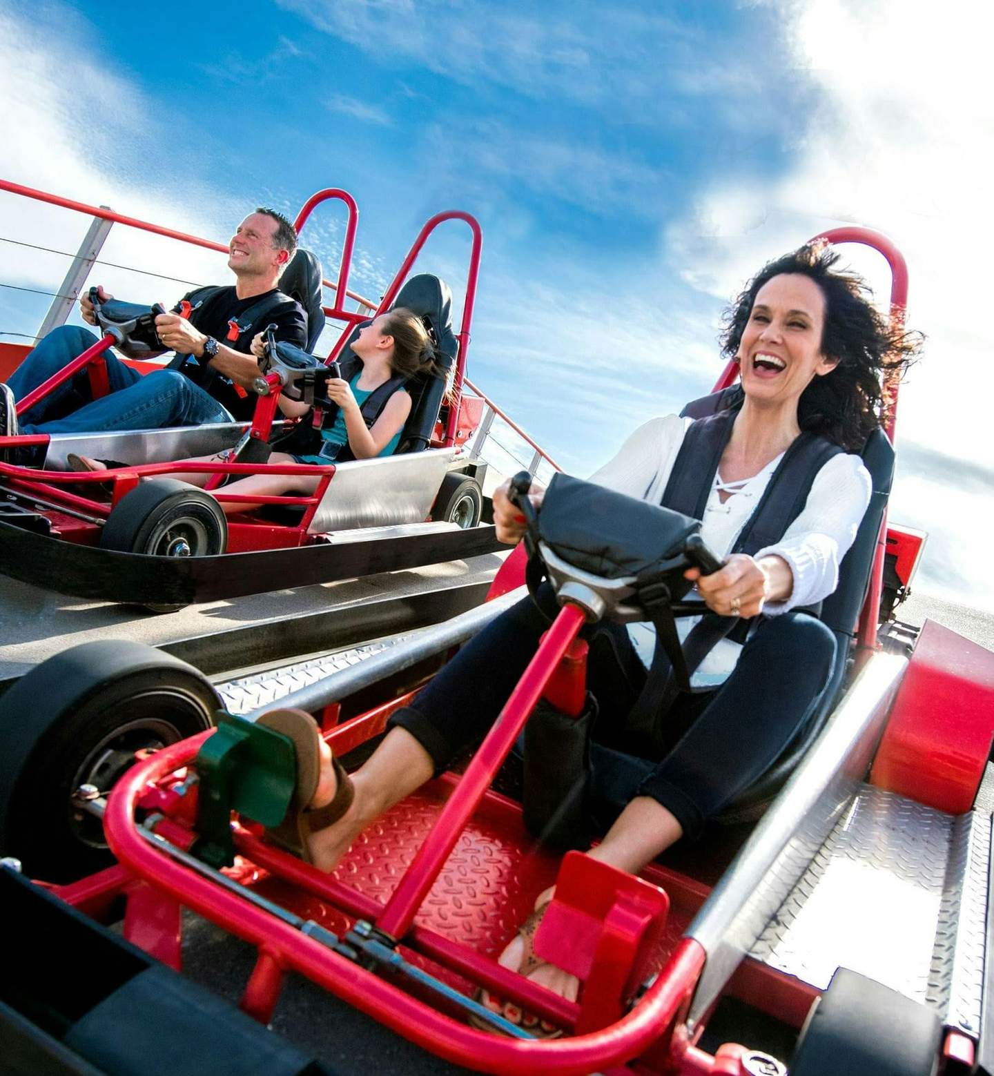 Two people in go-karts race on a track, smiling under a bright blue sky with some clouds. The scene is dynamic and joyful.