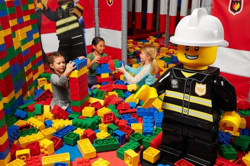 Three children play with large LEGO blocks in an indoor play area, featuring a life-sized LEGO firefighter figure.