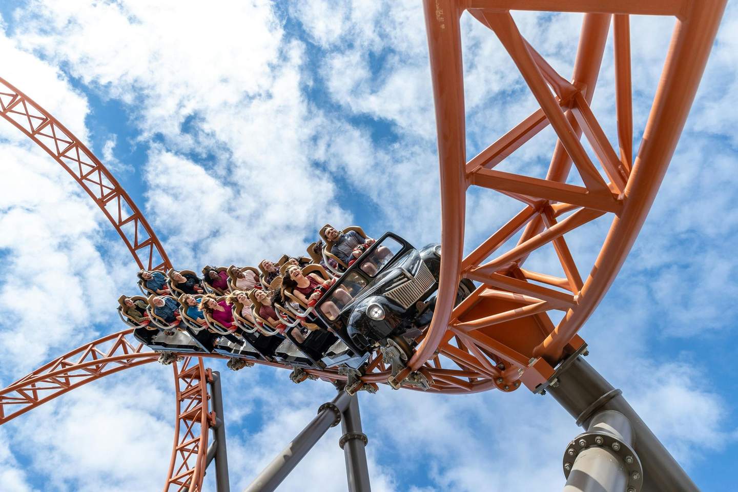 A roller coaster car full of people on orange tracks, captured against a blue sky with scattered clouds.