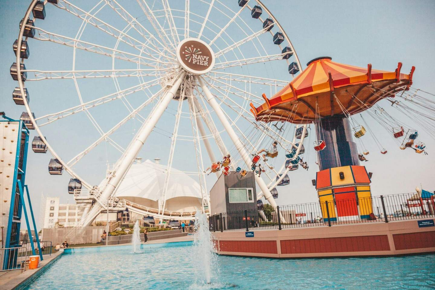 A Ferris wheel and a carousel with swings located near a pool with people around, all under a clear sky at Navy Pier.