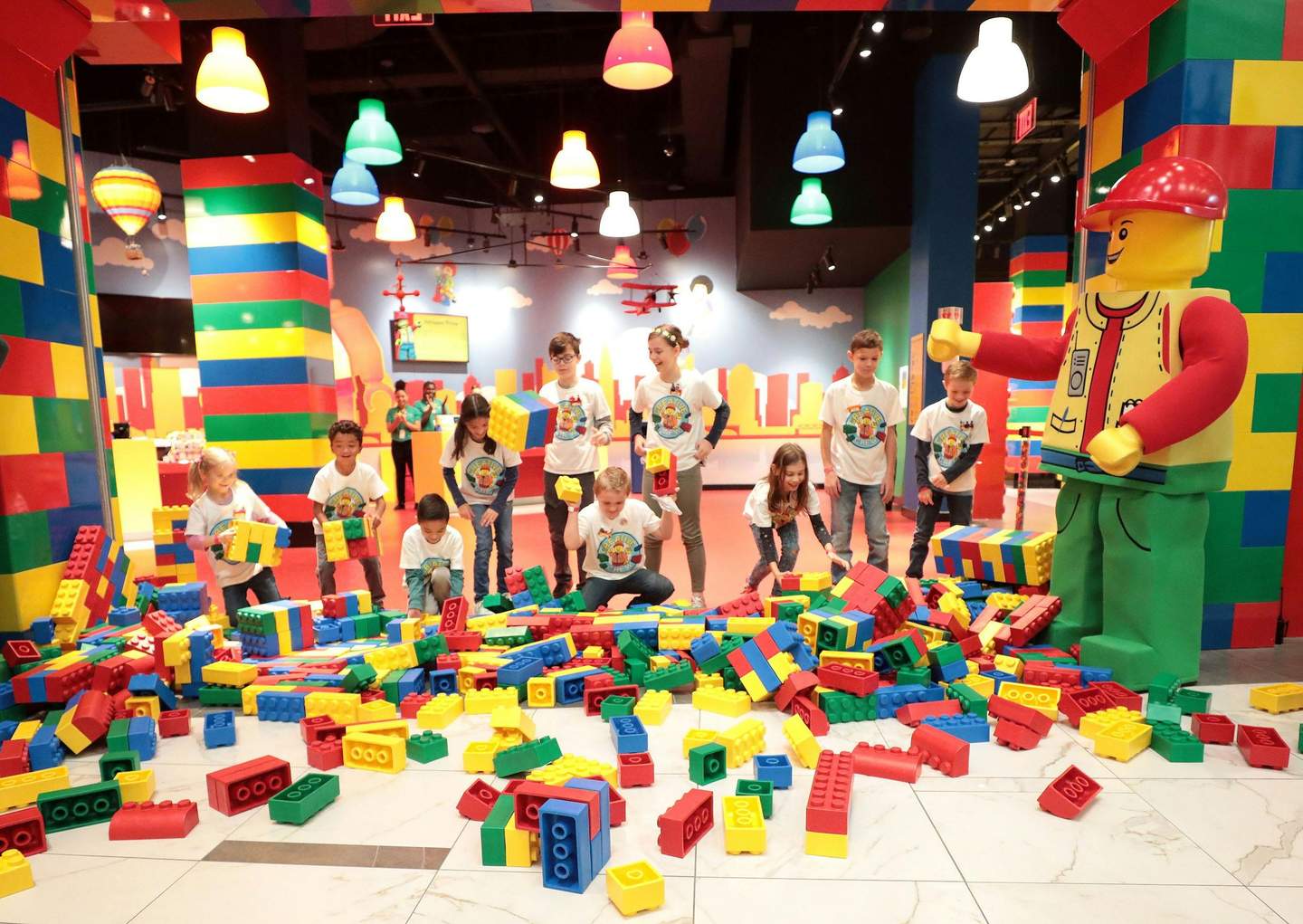 Children playing with large colorful toy bricks in a lively indoor space, surrounded by bright decorations and a giant LEGO figure.