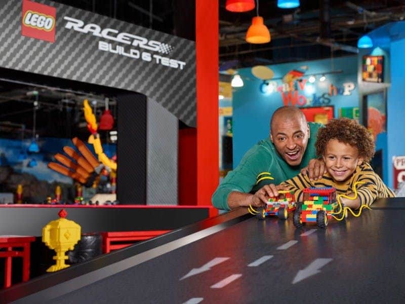 A man and child race LEGO cars on a sloped track in a colorful indoor play area with a sign reading "LEGO Racers Build & Test."