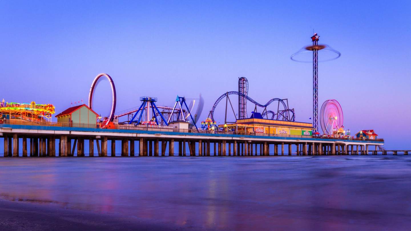 An amusement park pier with brightly lit rides, including a rollercoaster, Ferris wheel, and carousel, over calm water at dusk.
