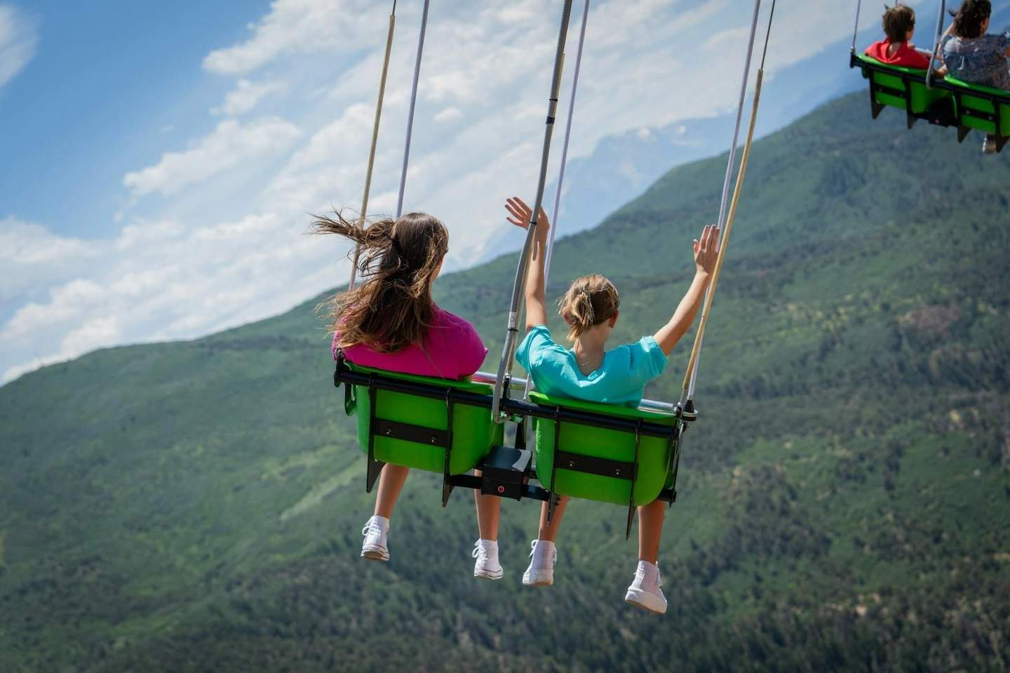Two people on a swing ride with their hands up, overlooking a scenic view of green mountains and a partly cloudy sky.