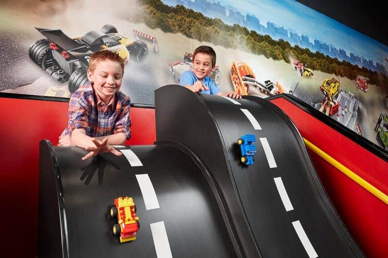 Two boys play with toy cars on black ramps featuring painted road markings, with colorful mural of racing cars in the background.