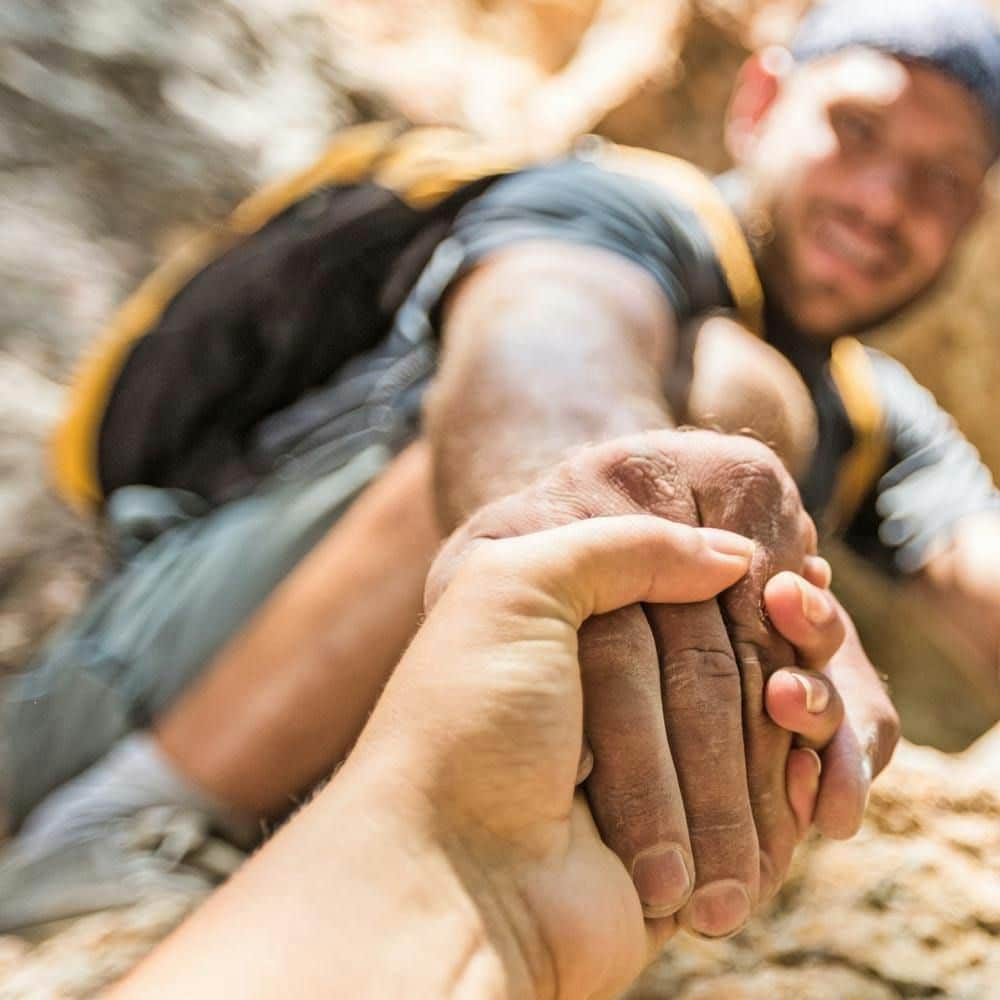A person helping another person up a rock formation by grasping their hand. The background appears to be rocky terrain.