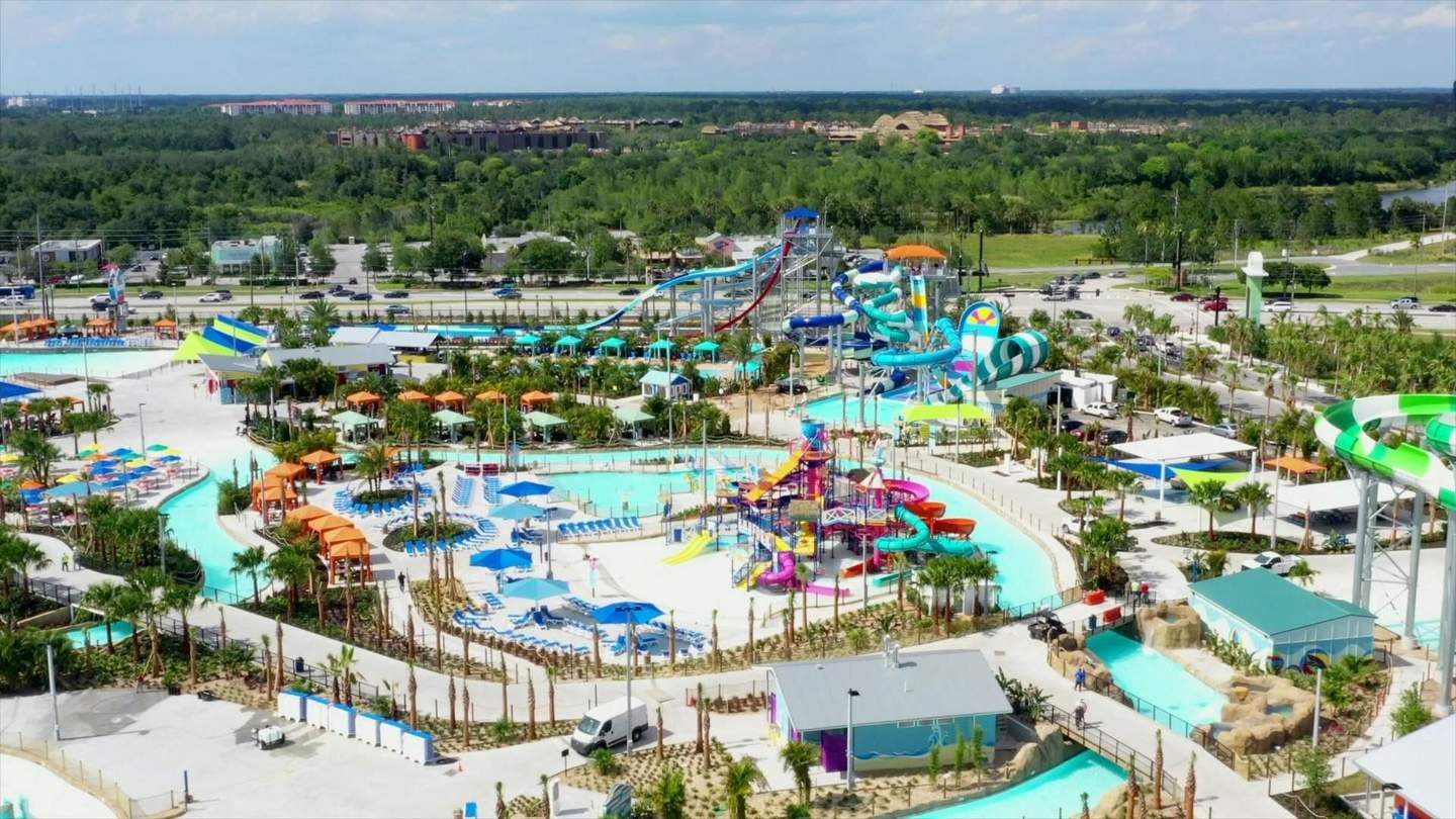 Aerial view of a large water park with various colorful slides, pools, lounge chairs, and shaded areas under blue skies.