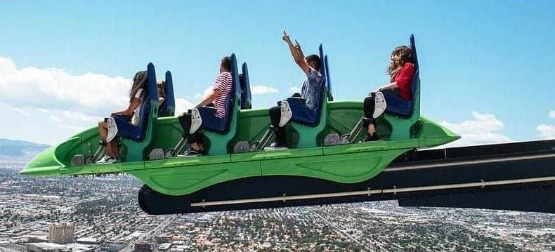 Four people on a green rollercoaster ride, suspended high above a cityscape under a blue sky with scattered clouds.