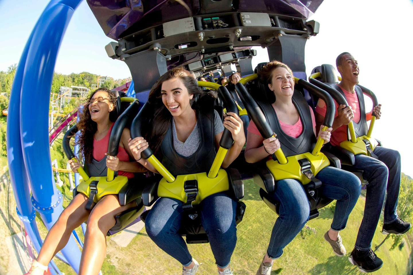 Four people smiling and laughing while riding a rollercoaster, seated in bright yellow and black harness seats.