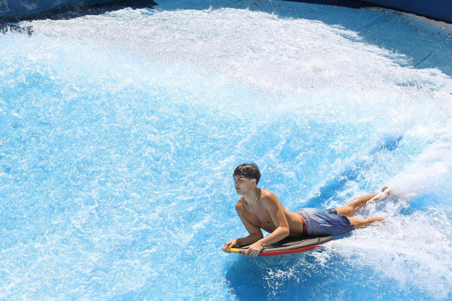 A person riding a bodyboard on a blue artificial wave at a water park.