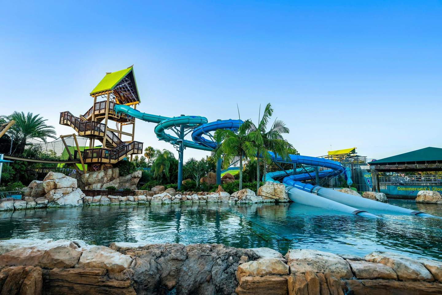 A tropical water park featuring a large, winding blue waterslide, palm trees, rocks, and a pool under a clear blue sky.