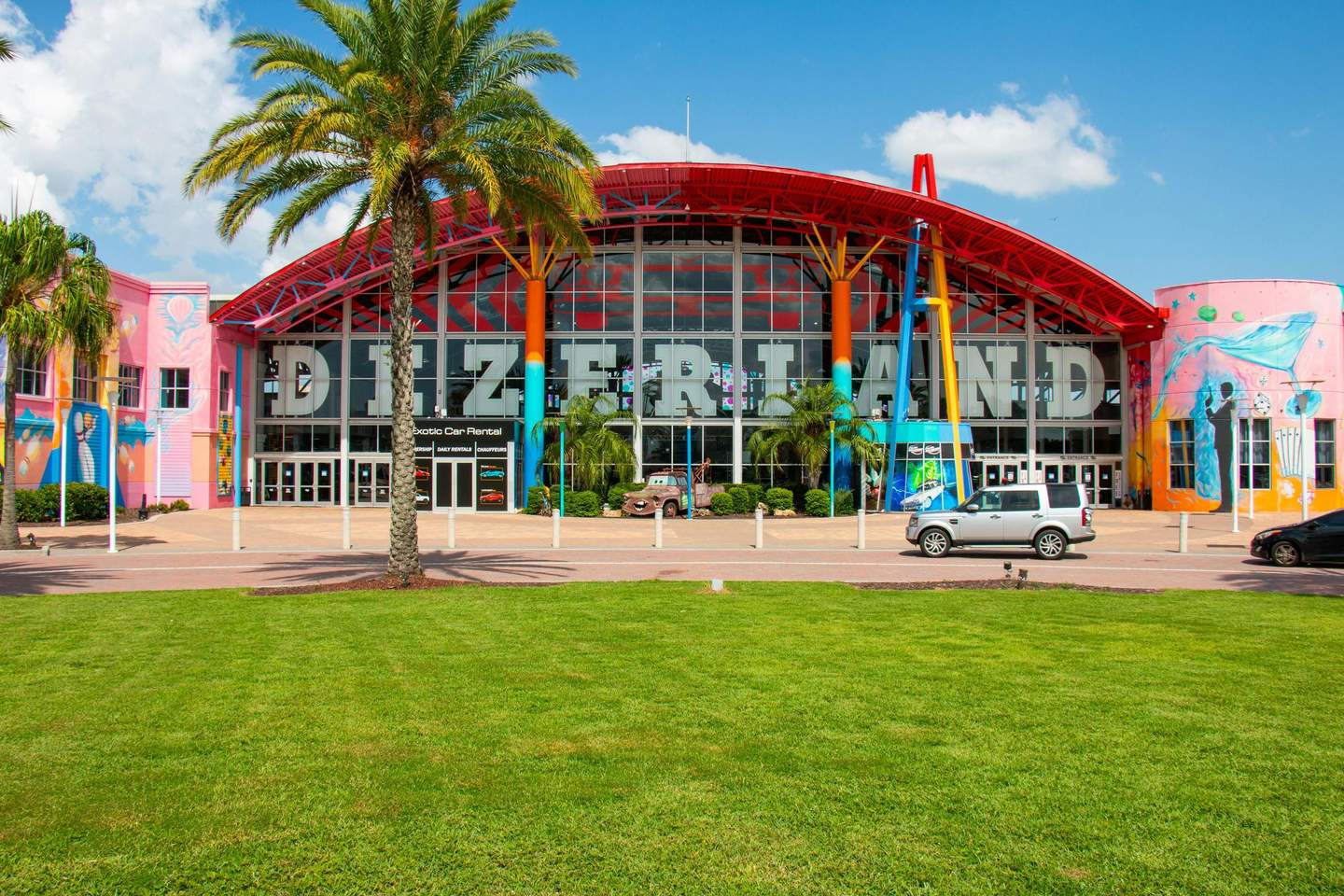 Dezerland Park Orlando Colorful building facade with the large word "DEZERLAND," flanked by palm trees, cars, and vibrant murals.