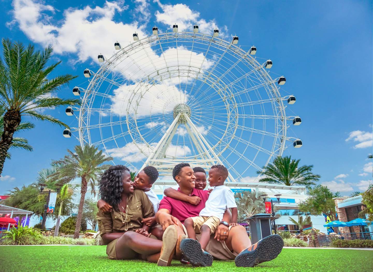 A family of five sits on grass, smiling in front of a large Ferris wheel, with palm trees and a clear blue sky in the background.
