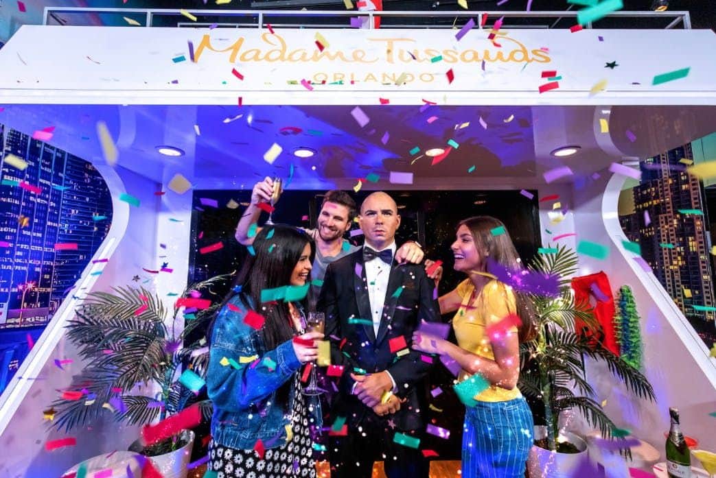 Three people celebrate with confetti and drinks around a wax figure in a tuxedo at Madame Tussauds Orlando.
