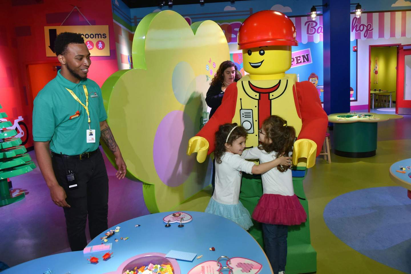 Two young girls hug a large Lego figure as a staff member smiles nearby in a colorful, toy-filled play area.