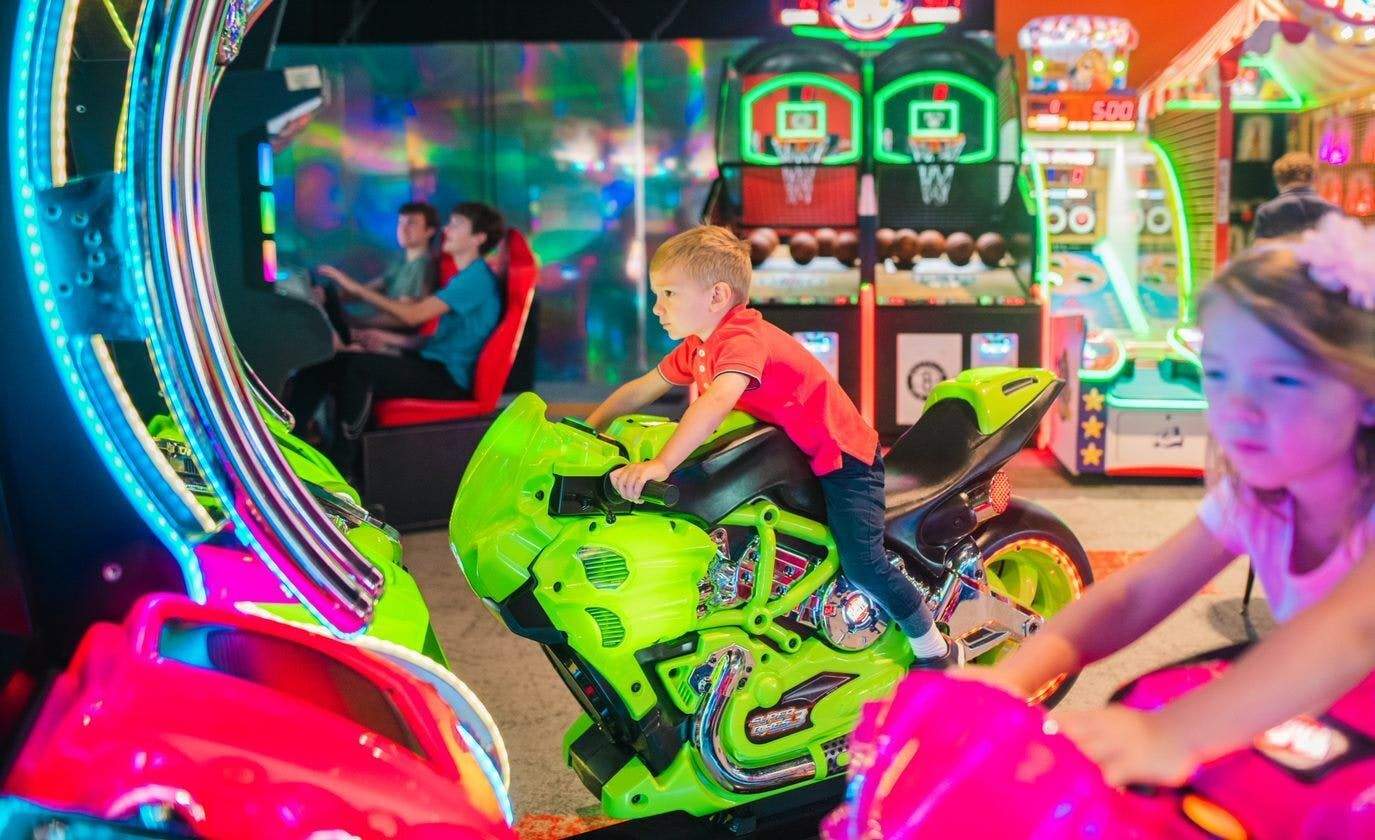 MagiQuest Unos niños están jugando en unos juegos arcade de motos, con un fondo colorido que incluye baloncesto y otras máquinas arcade.
