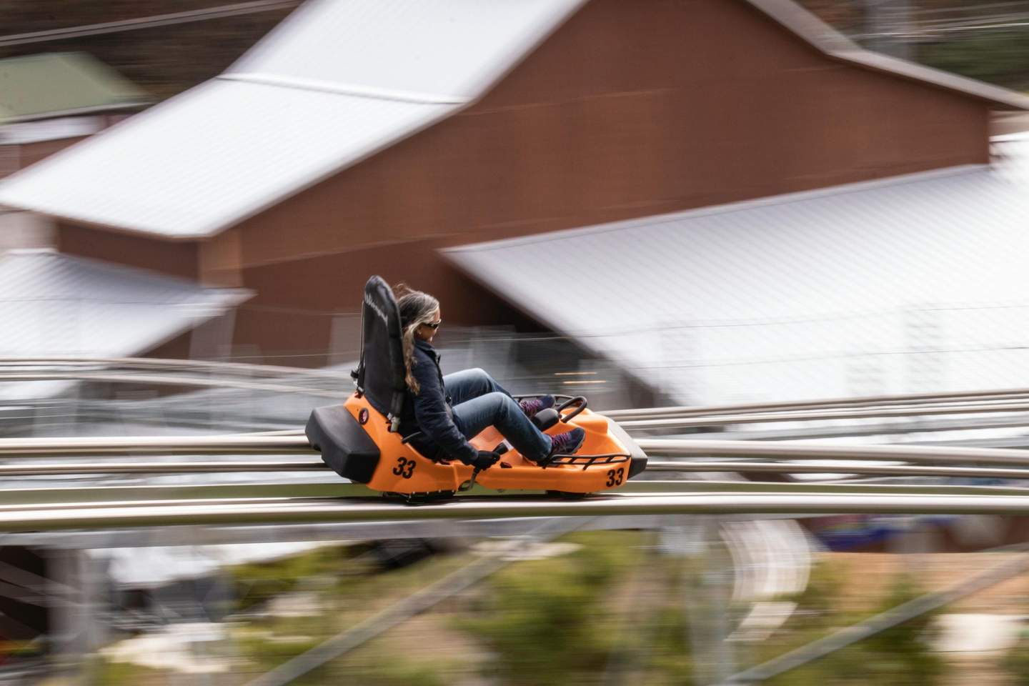Person riding an orange, single-seater roller coaster cart, moving swiftly against a blurred background of rooftops.