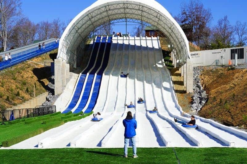 People sliding down multiple lanes of a tall, covered snow tubing slope; a person in a blue jacket watches from the bottom.